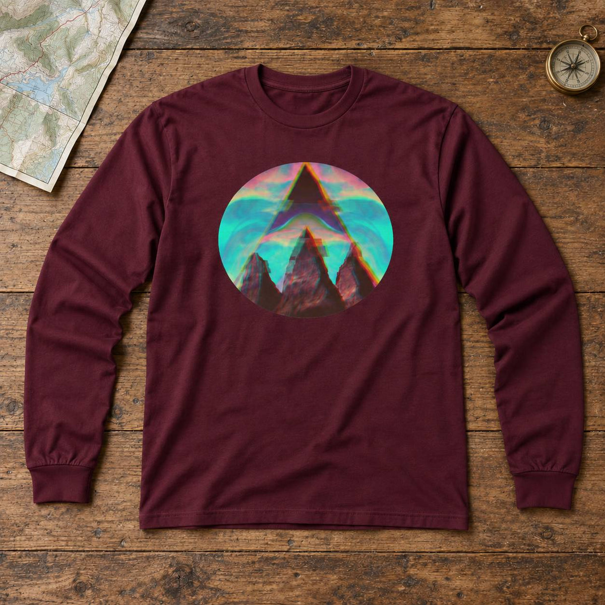 A long-sleeve crewneck t-shirt in maroon, laid flat on a rustic wooden surface. The shirt features a circular graphic print in the center showing a stylized mountain peak under a colorful sky with pink, orange, and teal tones.