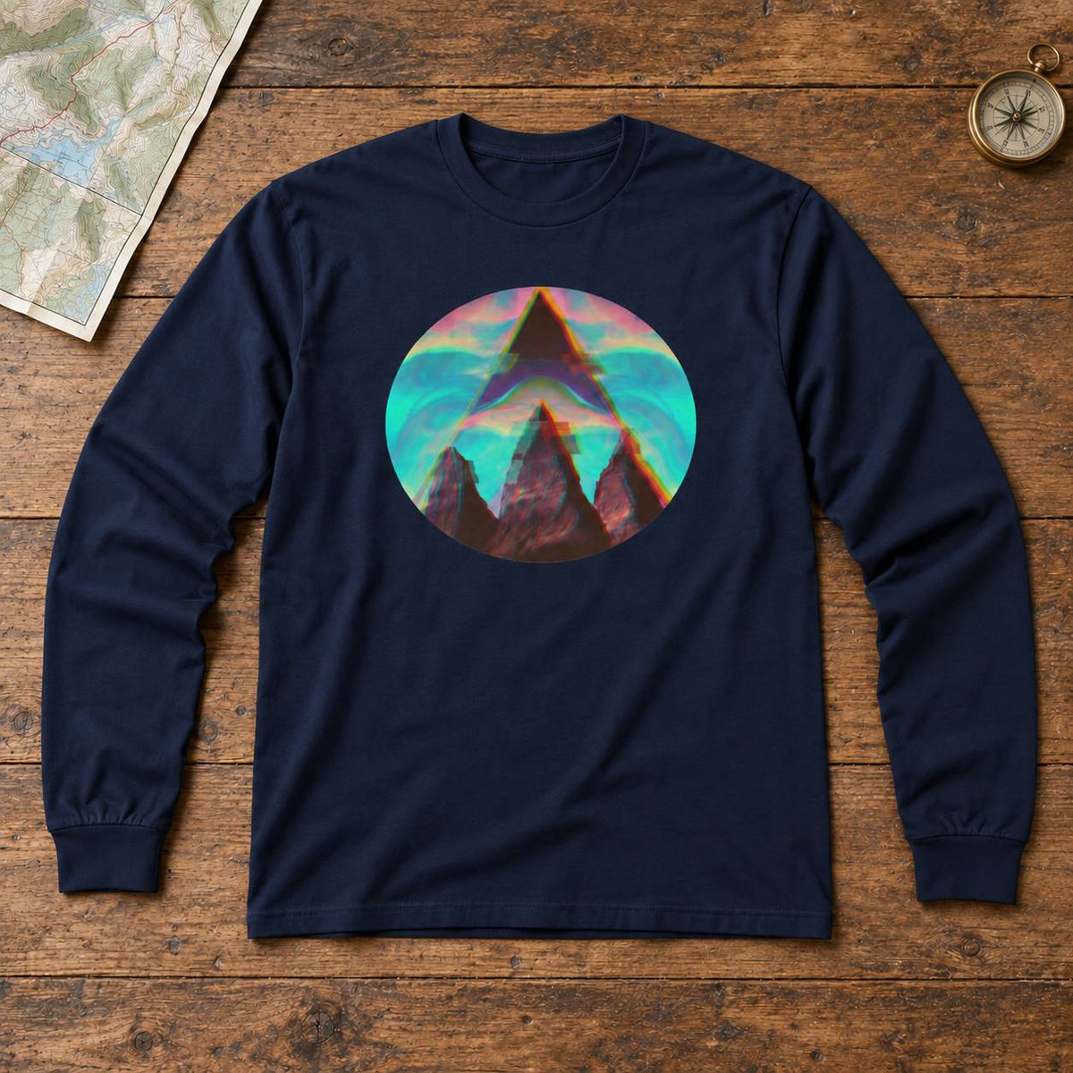 A navy blue long-sleeve t-shirt with a circular graphic print of a mountain peak in front of a colorful sunset, laid flat on a rustic wooden background with a map and compass as props.