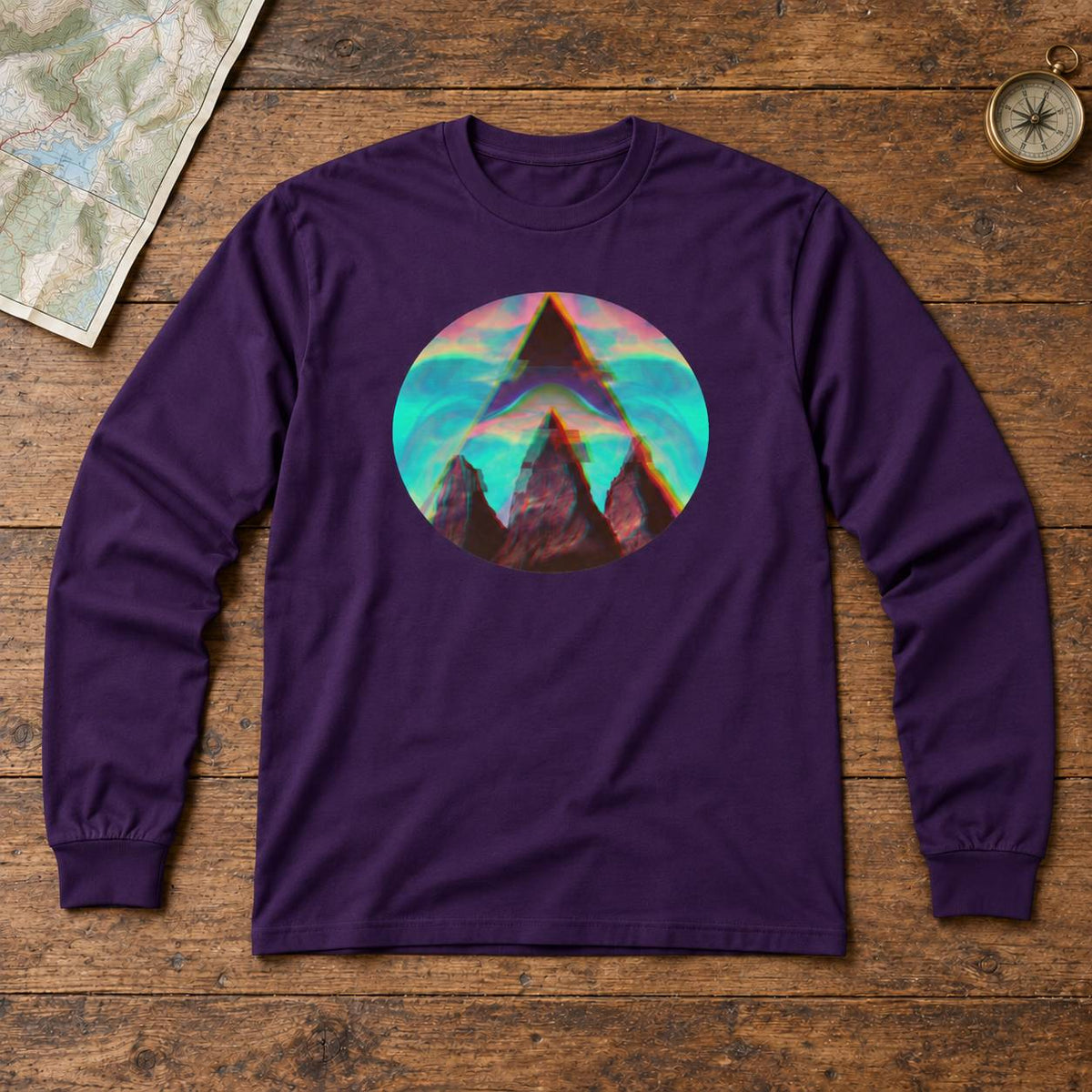 A purple long-sleeve t-shirt with a circular graphic print of a mountain peak in a colorful landscape, laid flat on a rustic wooden background with a map and compass as props.