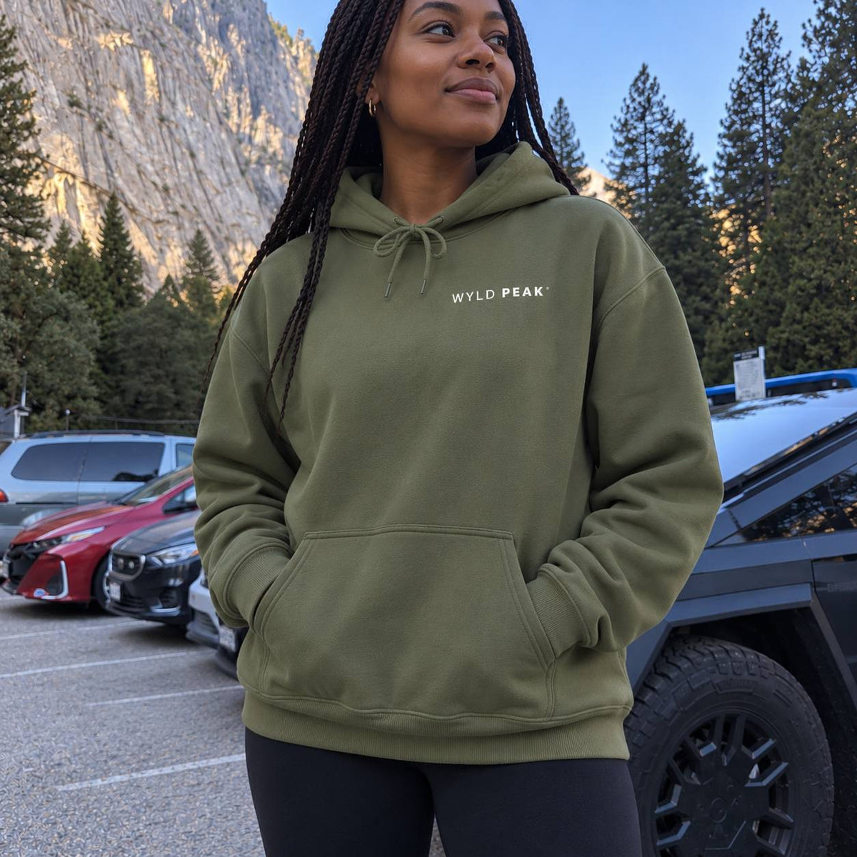 A woman wearing an olive green pullover hoodie with 'WYLD PEAK' printed in white on the chest, standing in a parking lot with mountains and trees in the background.