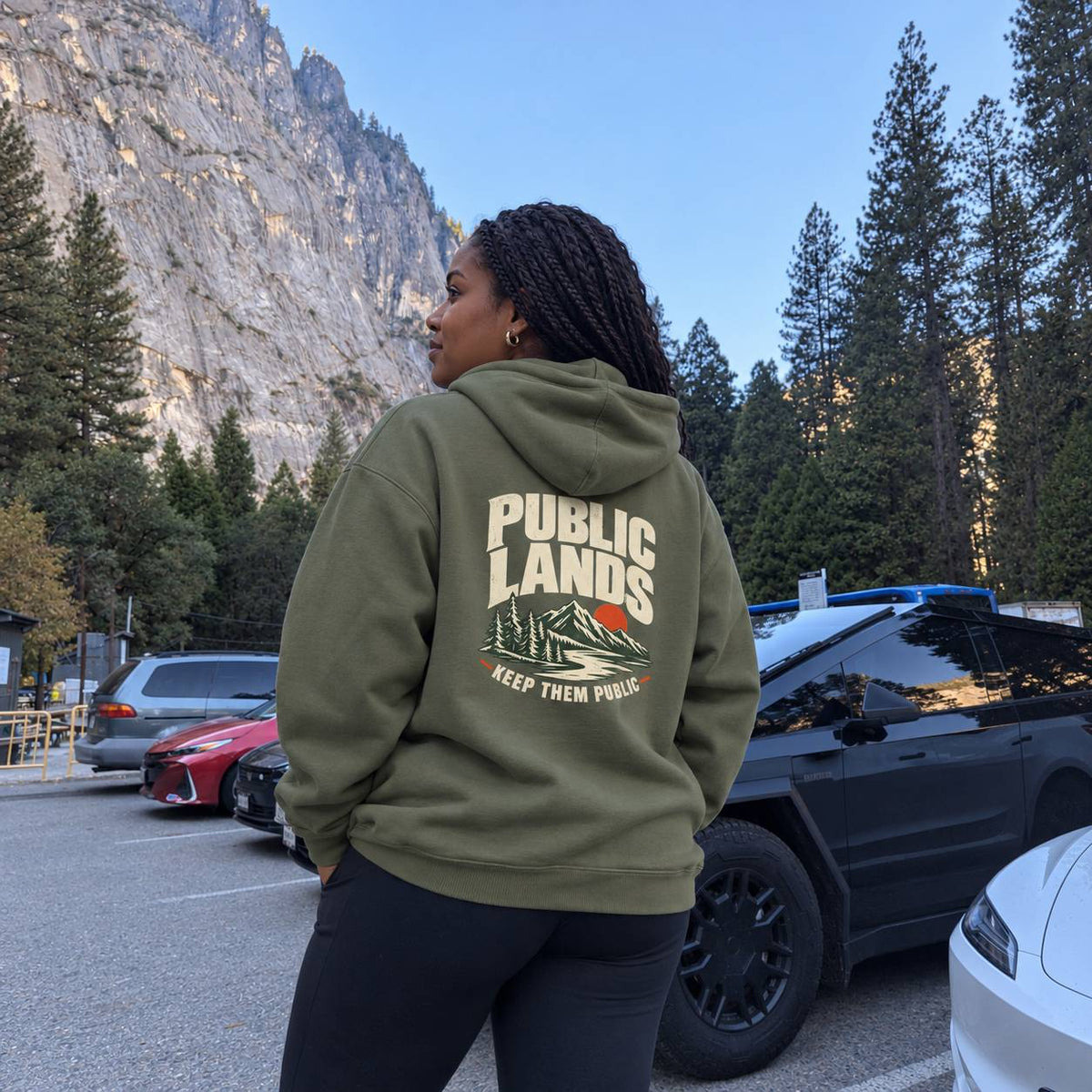 A woman stands in a parking lot outdoors, shown from the back wearing an olive green hoodie with a large graphic on the back that reads “PUBLIC LANDS” and “KEEP THEM PUBLIC.” Mountains, trees, and parked cars are visible in the background.