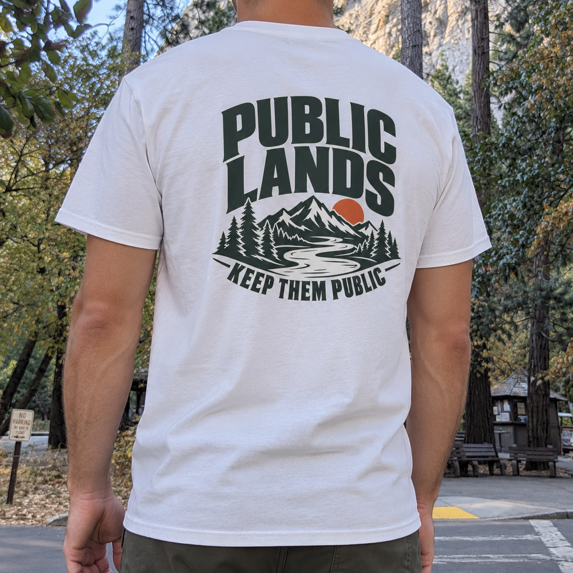 A man wearing a white t-shirt with a large back graphic that reads 'PUBLIC LANDS KEEP THEM PUBLIC' above a mountain, river, and pine tree illustration. The photo is taken outdoors in a park setting.