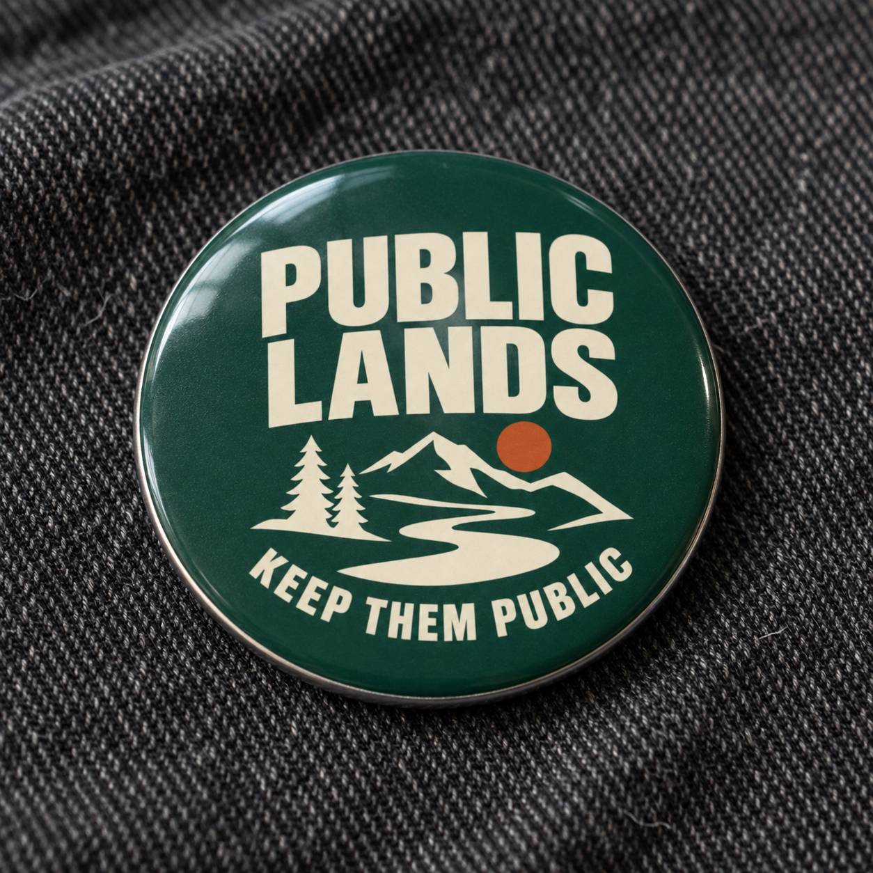 A round enamel pin with a dark green background and a silver metal rim, featuring the text 'PUBLIC LANDS' and 'KEEP THEM PUBLIC' in white, along with a white mountain, river, and tree graphic and a small orange sun. The pin is displayed on a dark gray textured fabric background.