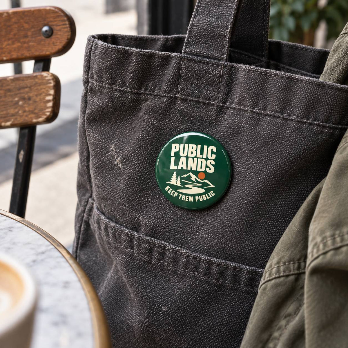A round green pin badge with white text reading 'PUBLIC LANDS' and 'KEEP THEM PUBLIC' with a mountain graphic, pinned to a dark canvas tote bag in an outdoor cafe setting.