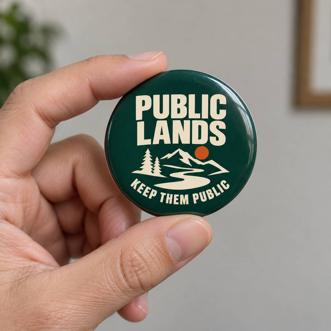 A hand holding a round dark green button badge with the text 'PUBLIC LANDS KEEP THEM PUBLIC' and a white mountain, river, and tree graphic with a small orange sun.