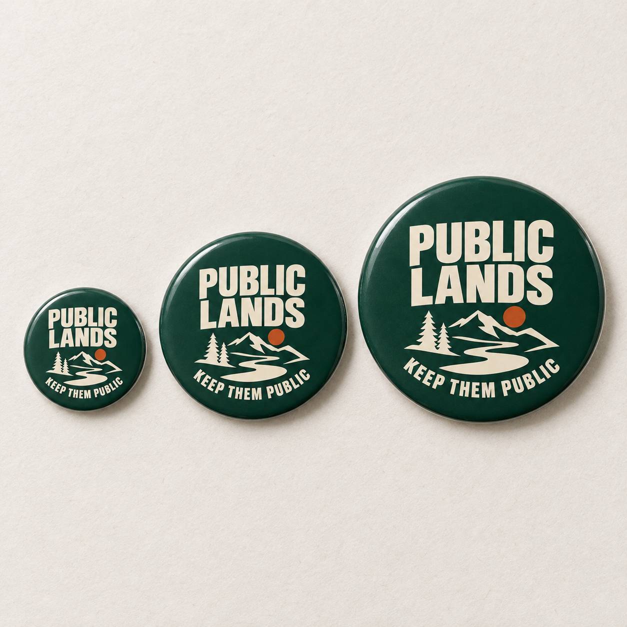 Three round pin-back buttons in dark green with a white mountain, river, and trees graphic, plus the words 'PUBLIC LANDS' and 'KEEP THEM PUBLIC' in white, displayed on a light beige background.
