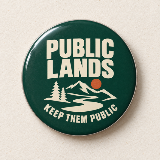 A round pin-back button badge with a dark green background and cream-colored text reading 'PUBLIC LANDS' and 'KEEP THEM PUBLIC,' plus a simple mountain, trees, and river graphic with a small orange sun. The badge is photographed on a light beige textured surface.