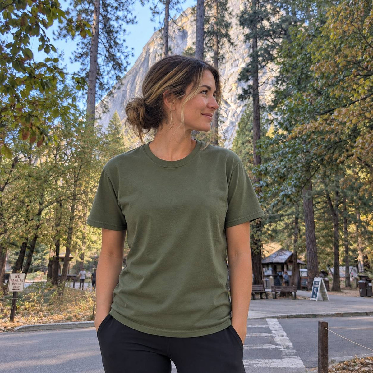 A woman wearing an olive green crewneck t-shirt and black pants, standing outdoors in a park with trees and a rocky cliff in the background.