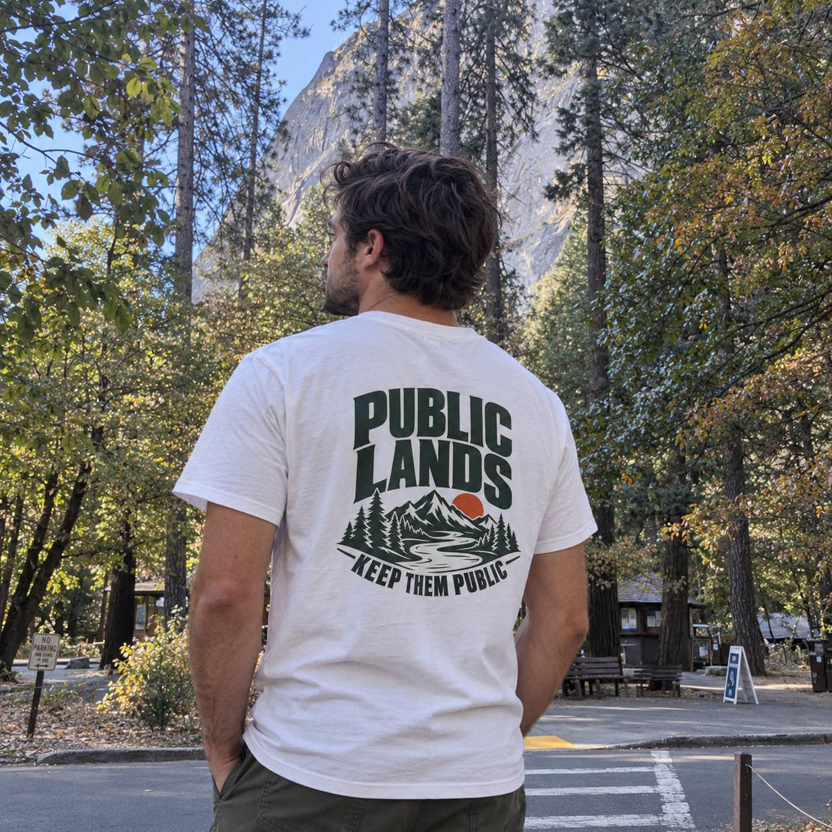A man viewed from behind wearing a white t-shirt with a green and orange graphic that reads 'PUBLIC LANDS KEEP THEM PUBLIC' with a mountain and trees illustration, standing in a forest park setting.