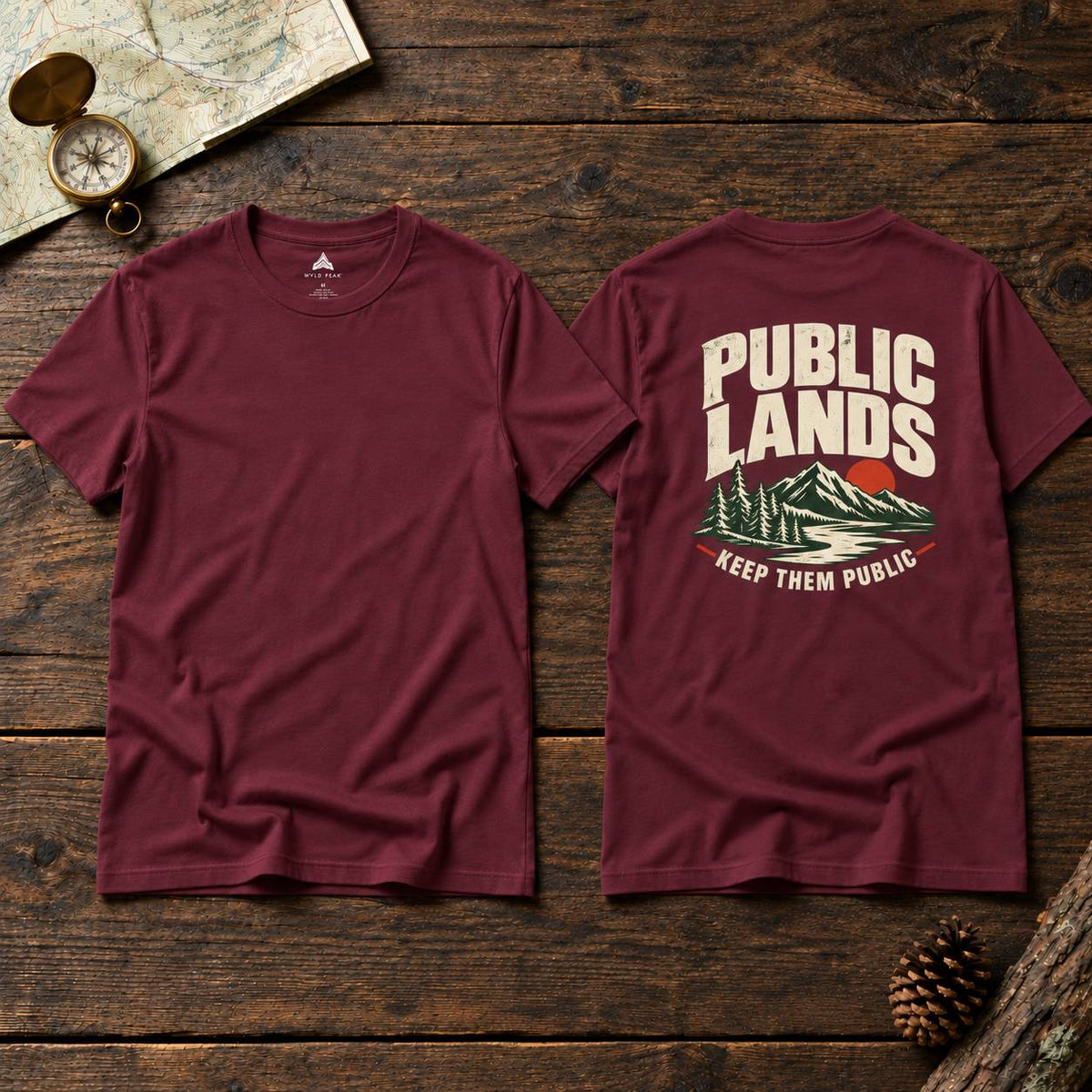 Two maroon short-sleeve t-shirts laid flat on a rustic dark wood surface, showing the front and back. The front has a small white chest logo, and the back has a large cream graphic reading 'PUBLIC LANDS' above a mountain-and-lake illustration with 'KEEP THEM PUBLIC' below.