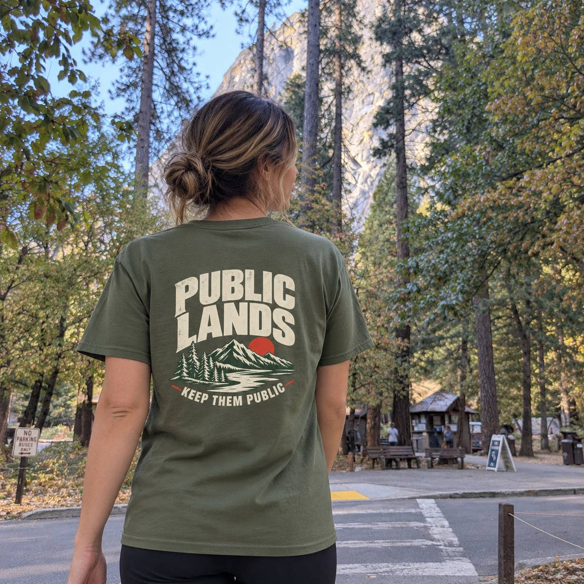 A woman viewed from behind wearing an olive green t-shirt with a graphic on the back that reads 'PUBLIC LANDS KEEP THEM PUBLIC' above a mountain and trees illustration. She is standing outdoors in a forested area with a rocky cliff in the background.