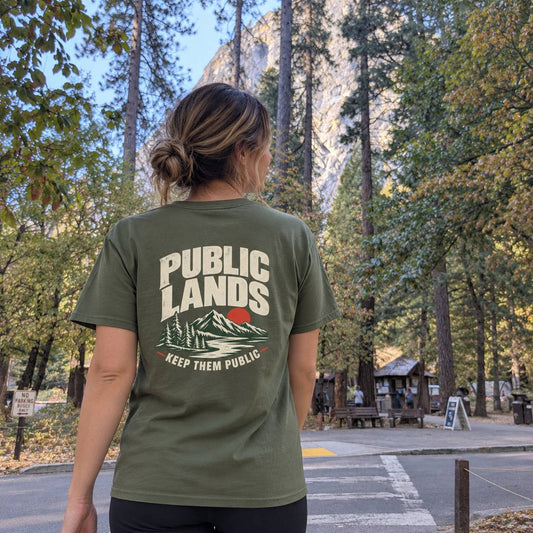 A woman viewed from behind wearing an olive green t-shirt with a graphic on the back that reads 'PUBLIC LANDS KEEP THEM PUBLIC' above a mountain and trees illustration. She is standing outdoors in a forested area with a rocky cliff in the background.