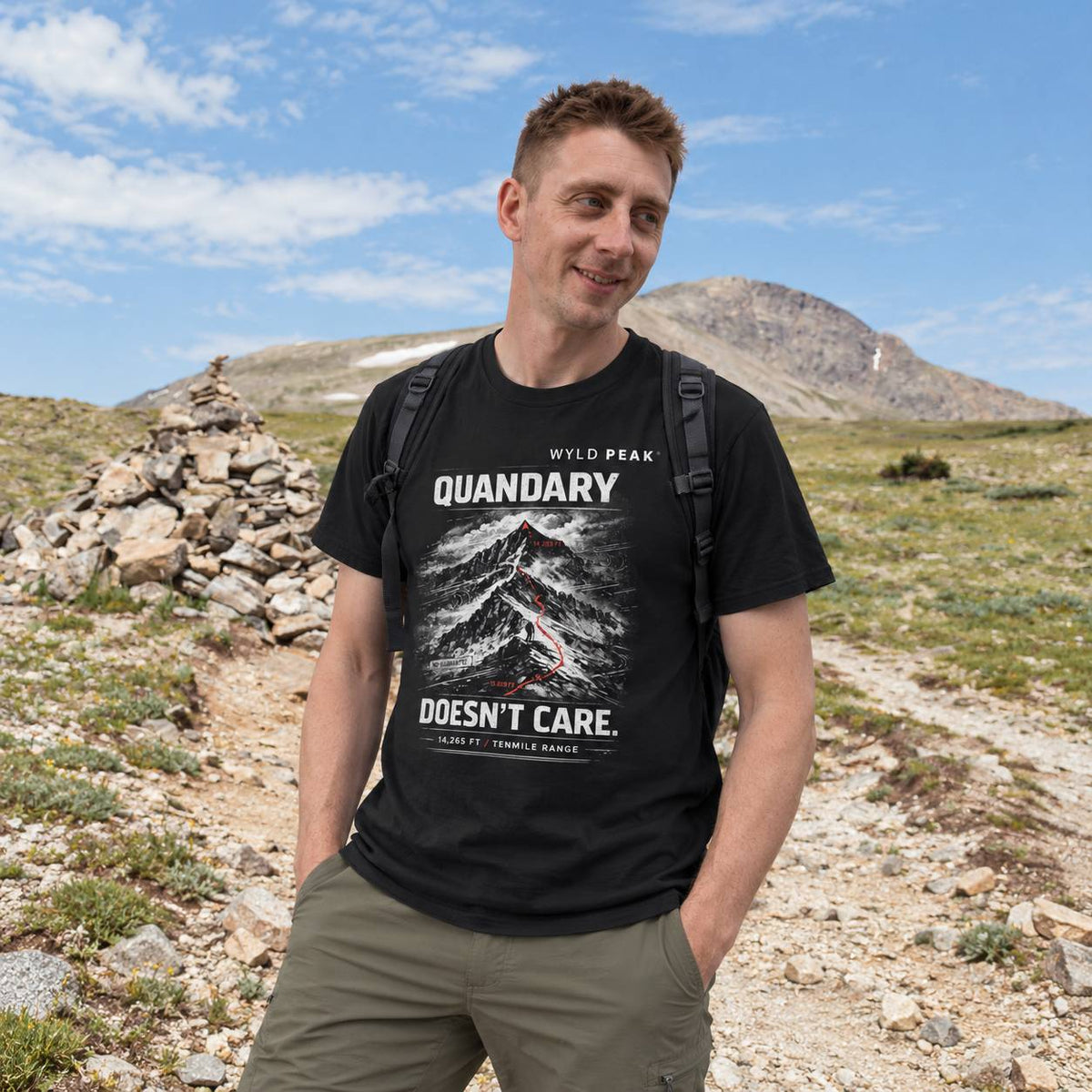 A man wearing a black graphic t-shirt with a mountain print and the text 'WYLD PEAK QUANDARY DOESN'T CARE.' stands outdoors in a mountain landscape.