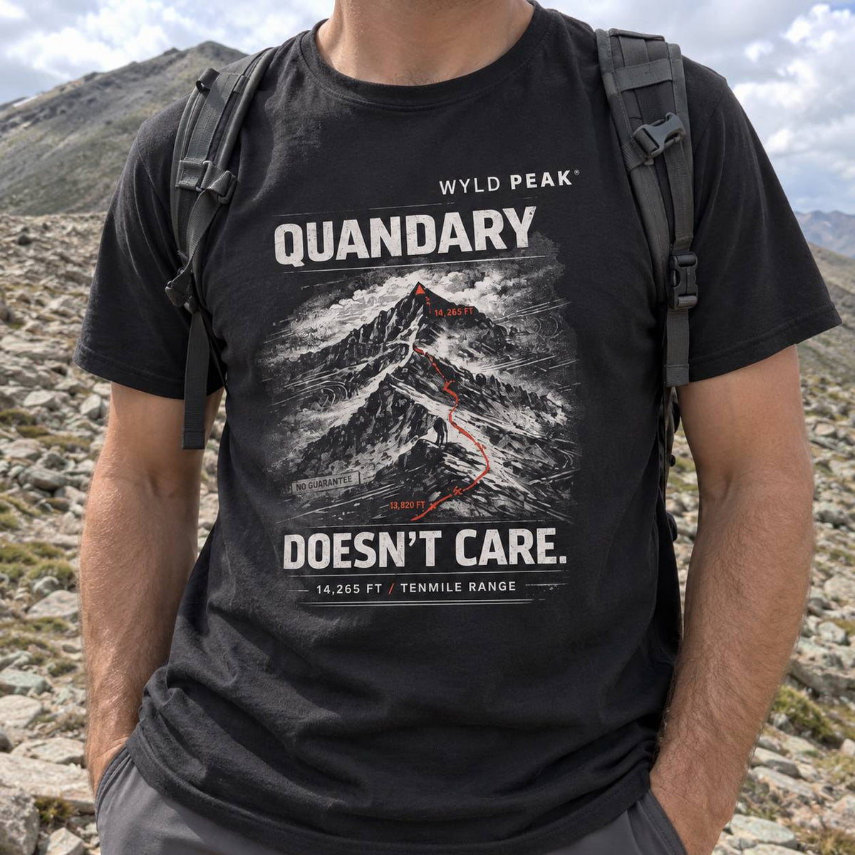 A man wearing a black graphic t-shirt with a mountain print and the text 'QUANDARY DOESN'T CARE.' in white and red, photographed outdoors in a rocky mountain landscape.