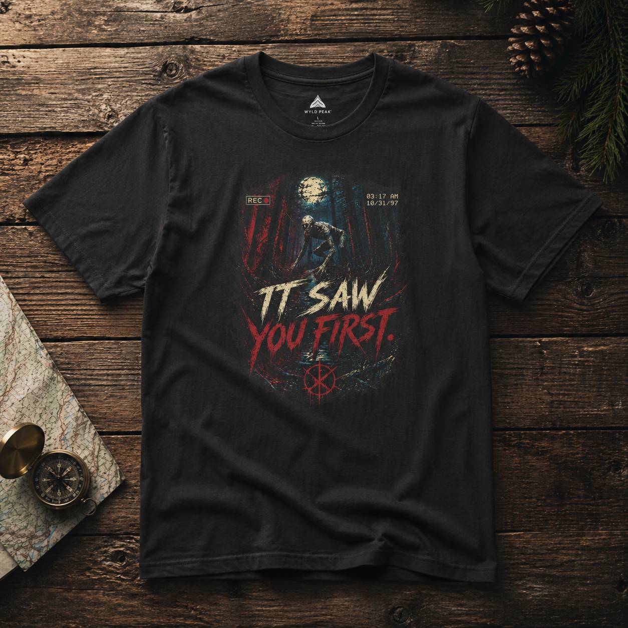 Black t-shirt with rake graphic design and text 'IT SAW YOU FIRST' on a wooden surface.