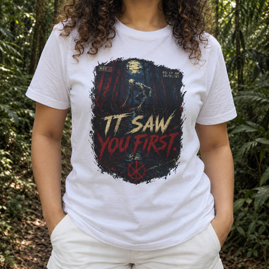 A person wearing a white graphic T-shirt with a dark horror-style print reading 'IT SAW YOU FIRST.' in red and yellow, photographed outdoors in a forest setting.