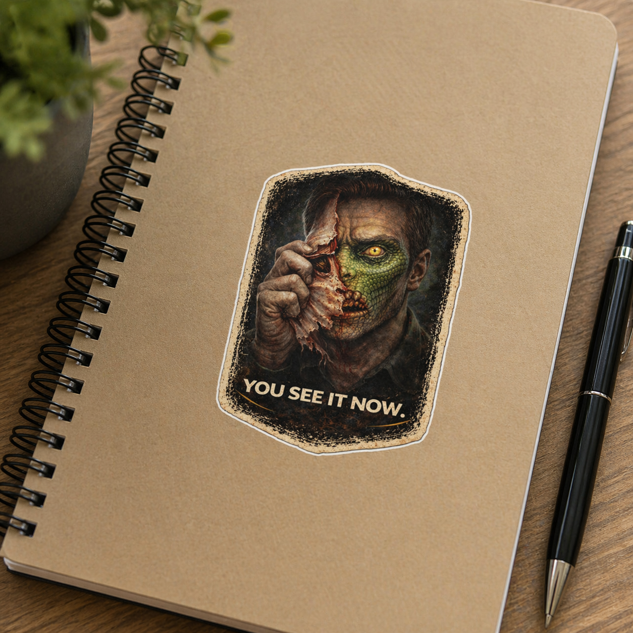 Notebook with a sticker of a character holding a severed head and the text 'You See It Now' on a wooden surface.