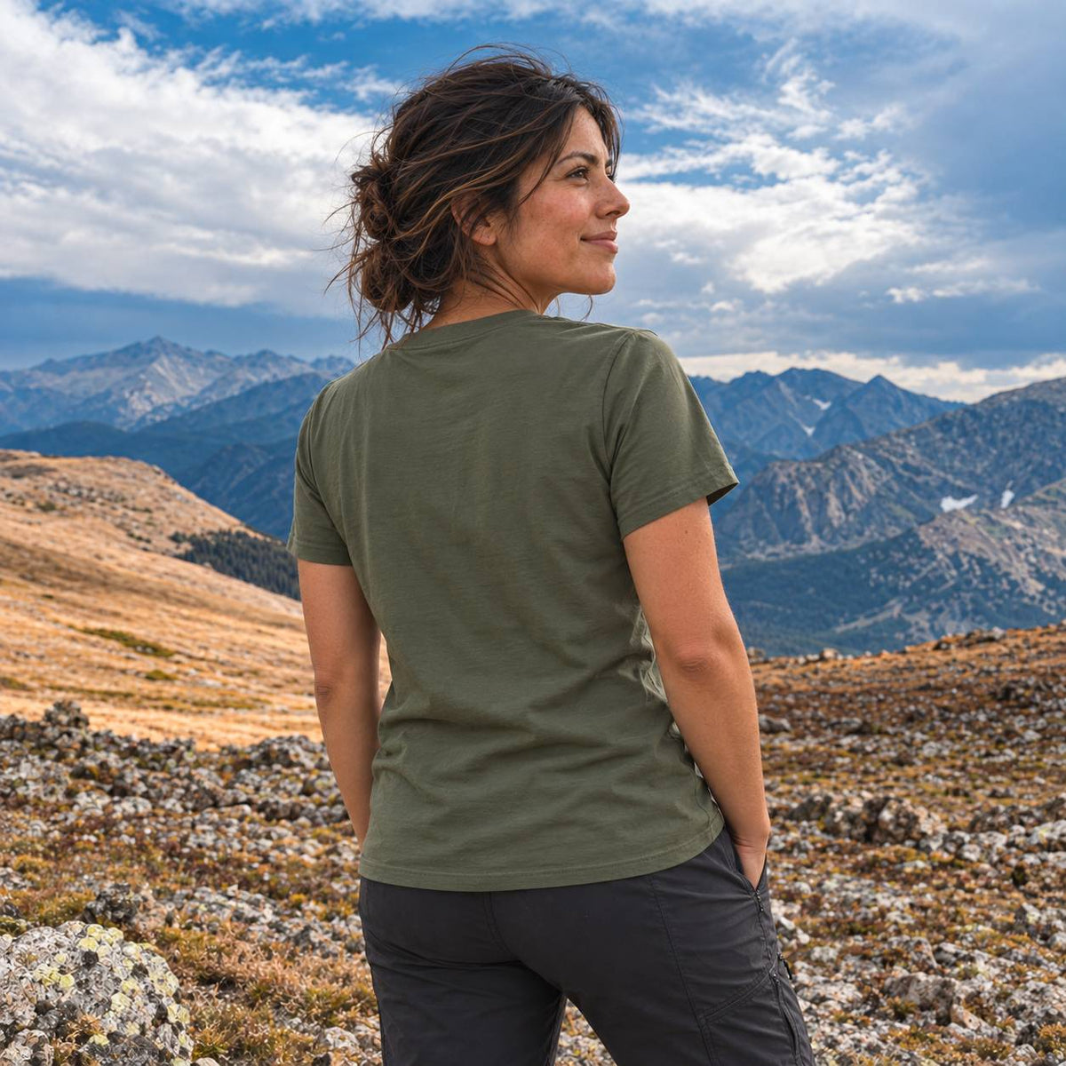 A woman wearing an olive green short-sleeve t-shirt and dark gray pants, photographed from behind as she looks out over a mountain landscape.