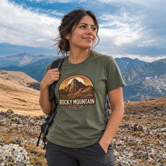 A woman hiking in a mountainous outdoor setting, wearing an olive green t-shirt with a circular 'Rocky Mountain' graphic on the chest and a black backpack.