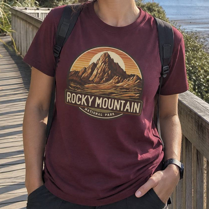 A person wearing a maroon t-shirt with a retro-style 'Rocky Mountain National Park' graphic, standing on a wooden boardwalk outdoors.