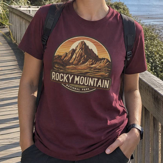 A person wearing a maroon t-shirt with a retro-style 'Rocky Mountain National Park' graphic, standing on a wooden boardwalk outdoors.