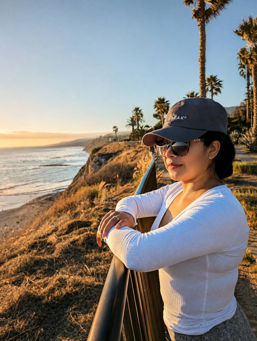 A woman wearing a dark gray baseball cap with 'Wyld Peak' printed on it, sunglasses, and a white ribbed long-sleeve top leans on a railing at a coastal cliffside location during golden hour.