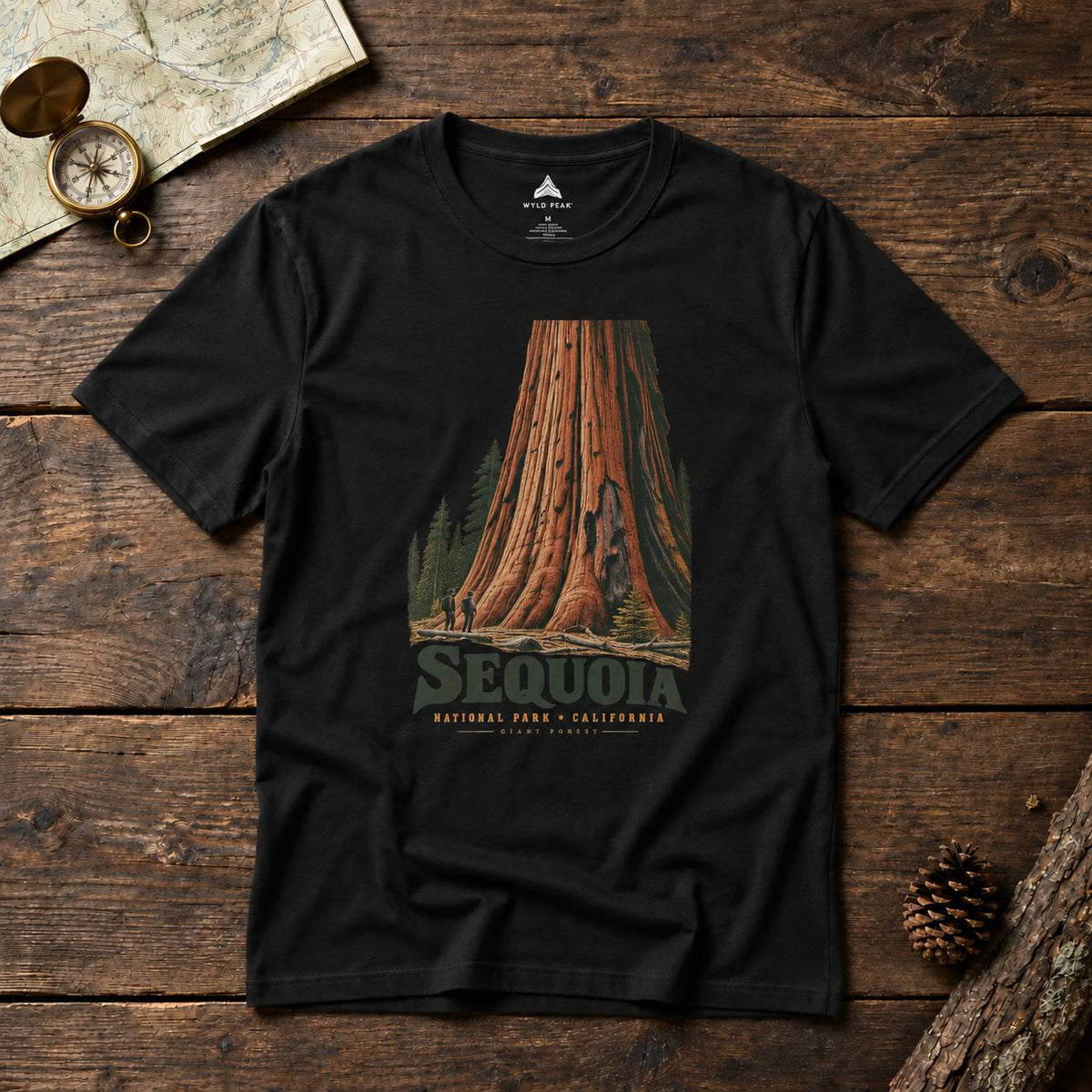 A black short-sleeve graphic t-shirt featuring a vintage-style illustration of a giant sequoia tree with 'SEQUOIA NATURAL PARK - CALIFORNIA' printed below it. The shirt is displayed flat on a rustic dark wood background alongside a map, compass, and pinecone.
