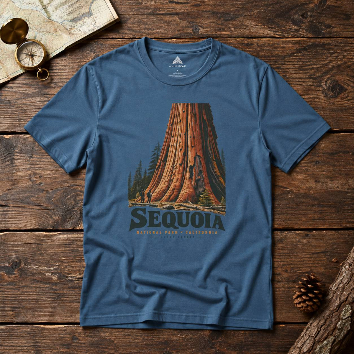 A blue short-sleeve t-shirt with a vintage-style graphic of a giant sequoia tree and the text 'SEQUOIA NATIONAL PARK, CALIFORNIA' laid flat on a rustic wooden surface with a map, compass, and pinecone props.