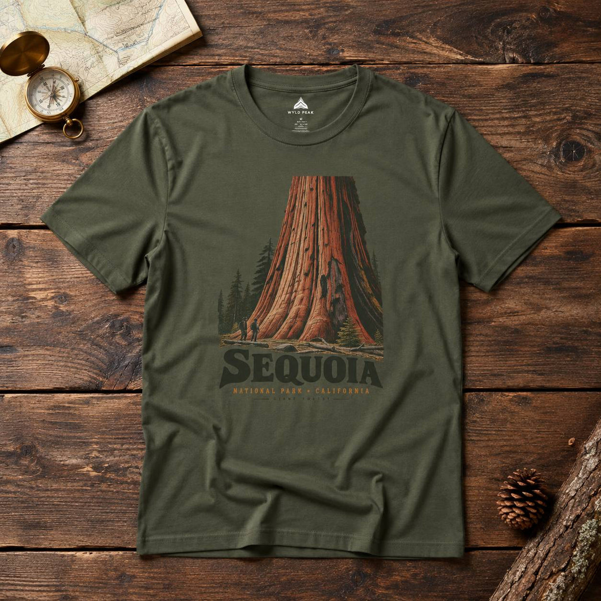 An olive green short-sleeve t-shirt with a vintage-style redwood tree graphic and the word 'SEQUOIA' printed on the front, laid flat on a rustic dark wood background with a map, compass, and pinecone nearby.
