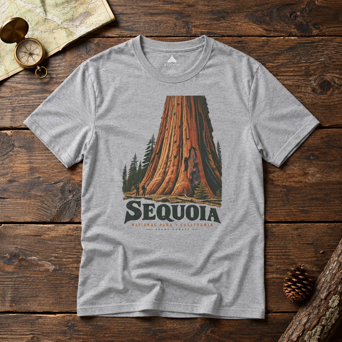 A heather gray short-sleeve t-shirt with a vintage-style Sequoia National Park graphic, laid flat on a rustic dark wood background with a map, compass, and pinecone props.