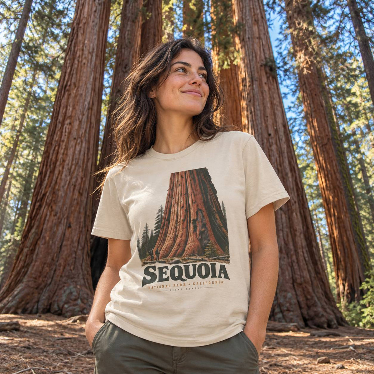 A woman wearing a beige graphic t-shirt with a Sequoia redwood tree print, standing in a forest of giant sequoia trees.
