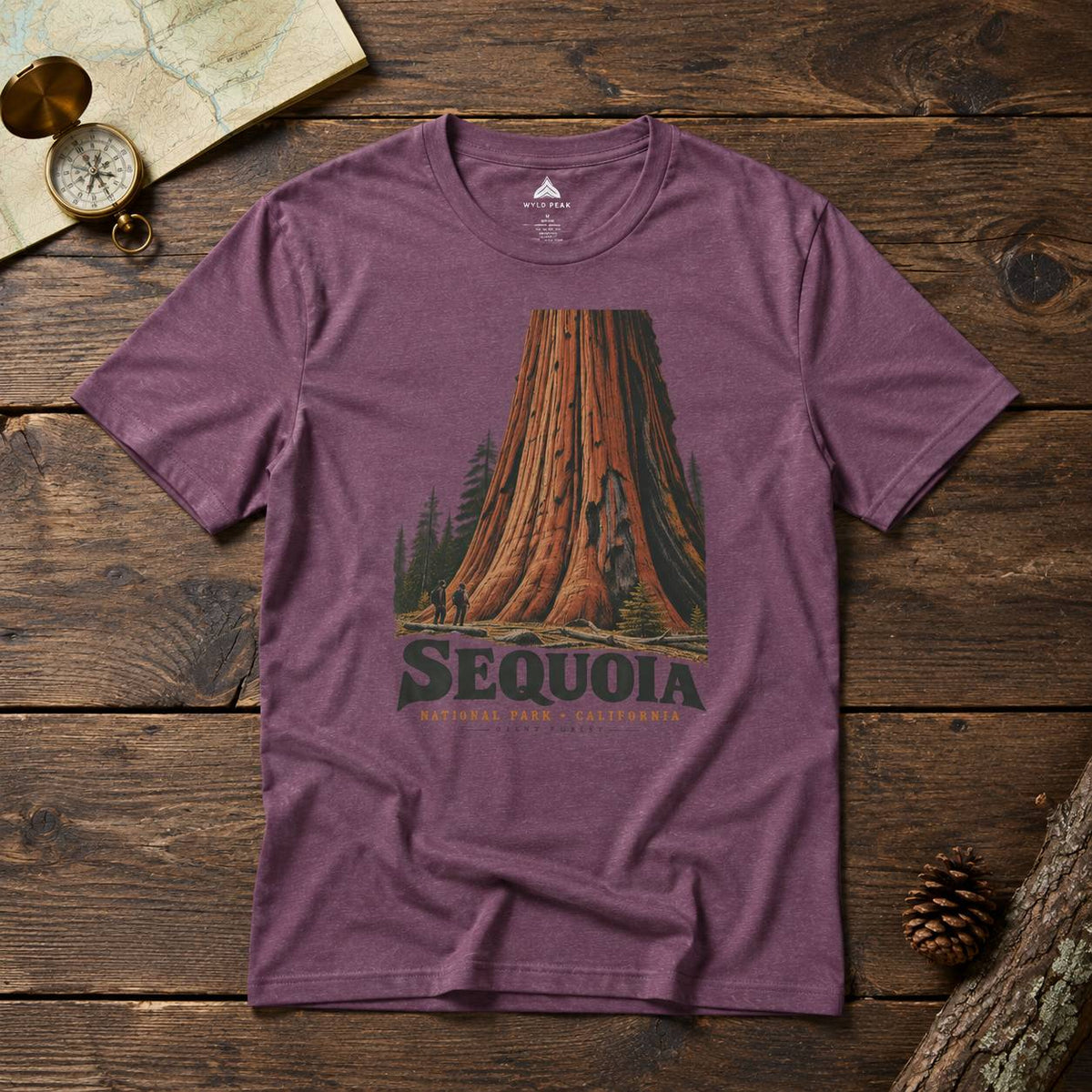A heathered purple short-sleeve t-shirt featuring a vintage-style graphic of a giant sequoia tree with the word 'SEQUOIA' printed below it. The shirt is laid flat on a rustic dark wood background, surrounded by decorative props including a vintage compass, a map, and a pinecone.