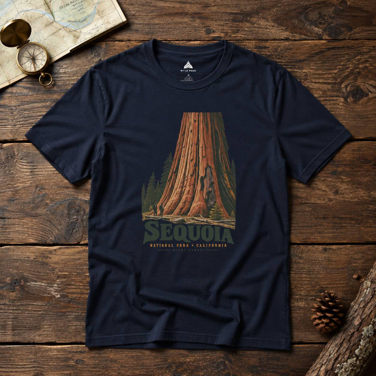 A navy blue crewneck t-shirt featuring a vintage-style graphic of a giant sequoia tree with the text 'SEQUOIA' and 'NATURAL WILD CALIFORNIA' printed on the front. The shirt is displayed in a flat-lay arrangement on a rustic wooden surface, surrounded by a map, a brass compass, and a pinecone.