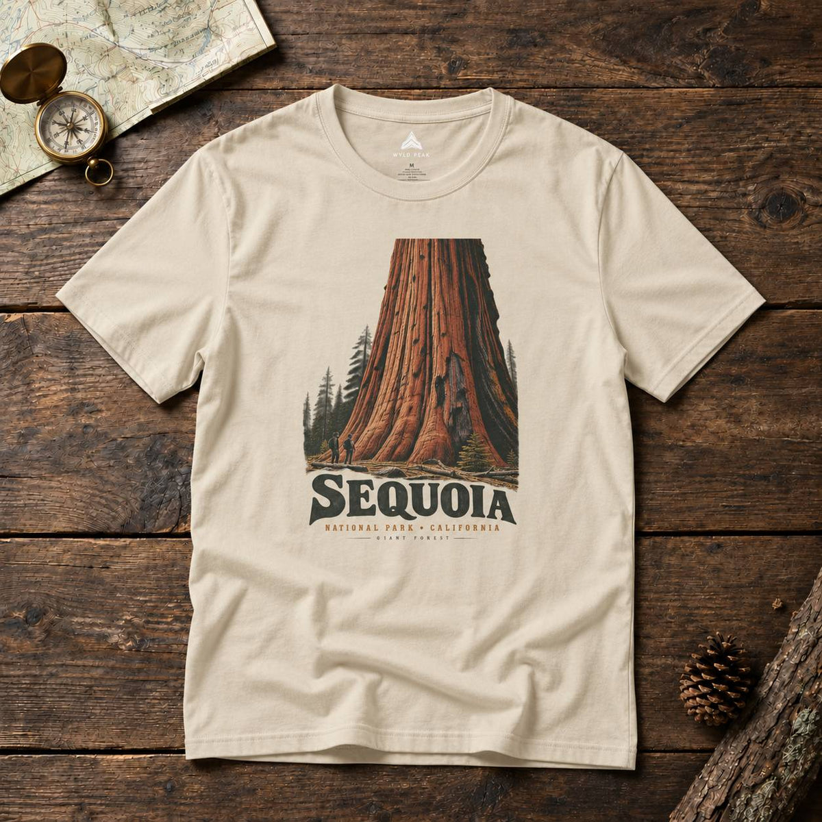 A beige crewneck t-shirt featuring a vintage-style graphic of a giant sequoia tree and the text 'SEQUOIA NATURAL PARK, CALIFORNIA' laid flat on a rustic dark wood background with outdoor props.