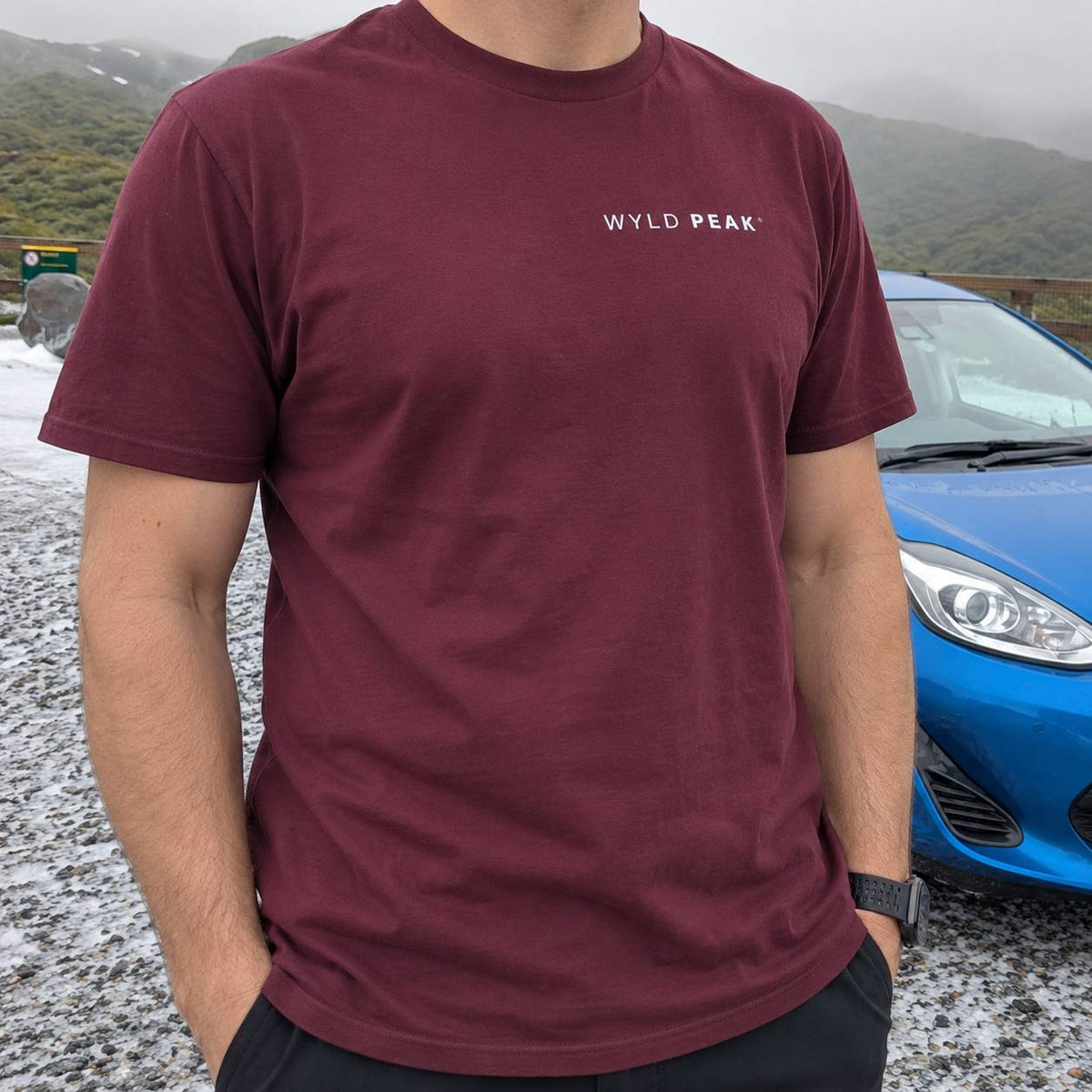 A man wearing a maroon crewneck t-shirt with 'WYLD PEAK.' printed in white on the chest, standing outdoors next to a blue car with mountains in the background.