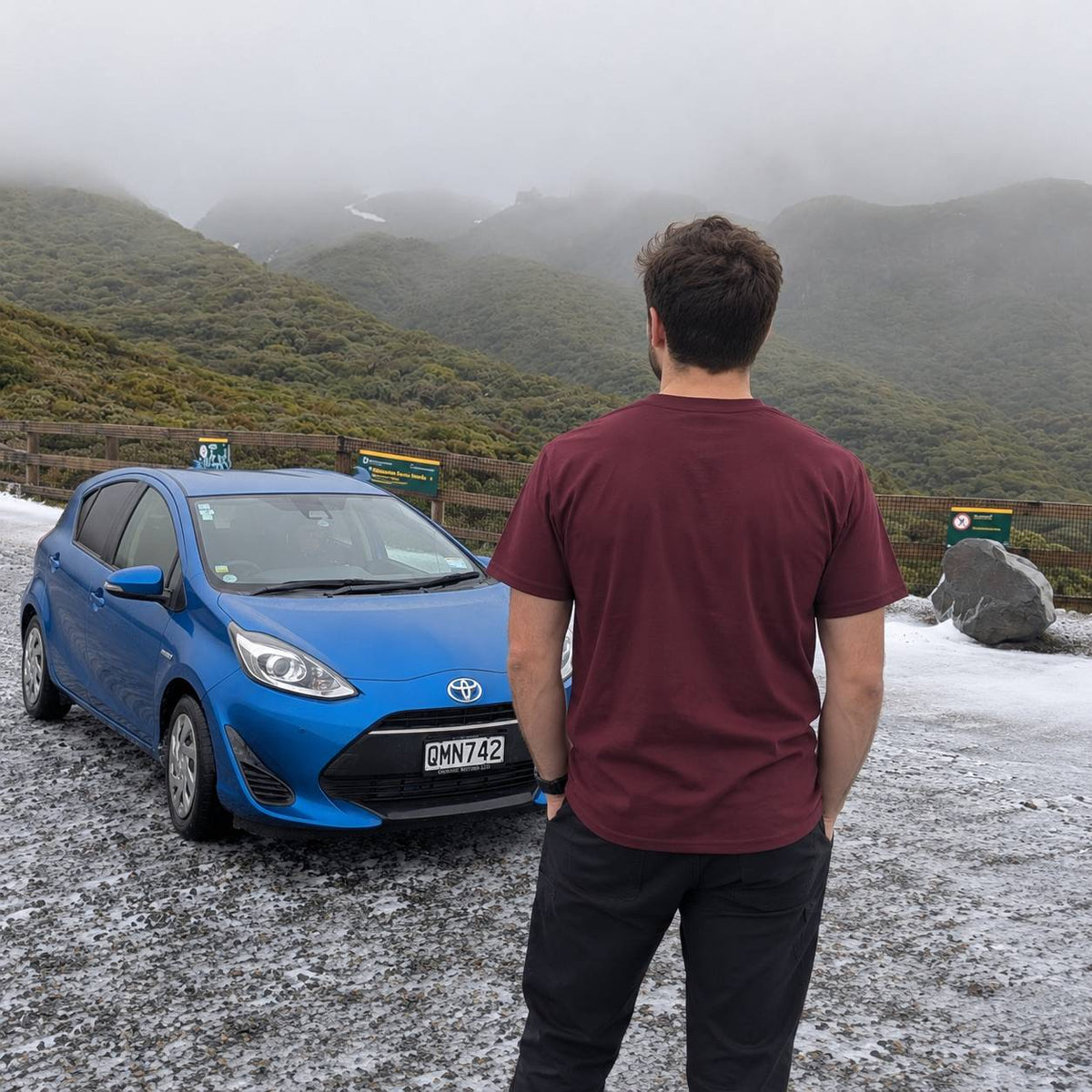 A man in a maroon t-shirt and black pants stands on a snowy mountain road facing misty green hills, with a blue Toyota hatchback parked next to him.
