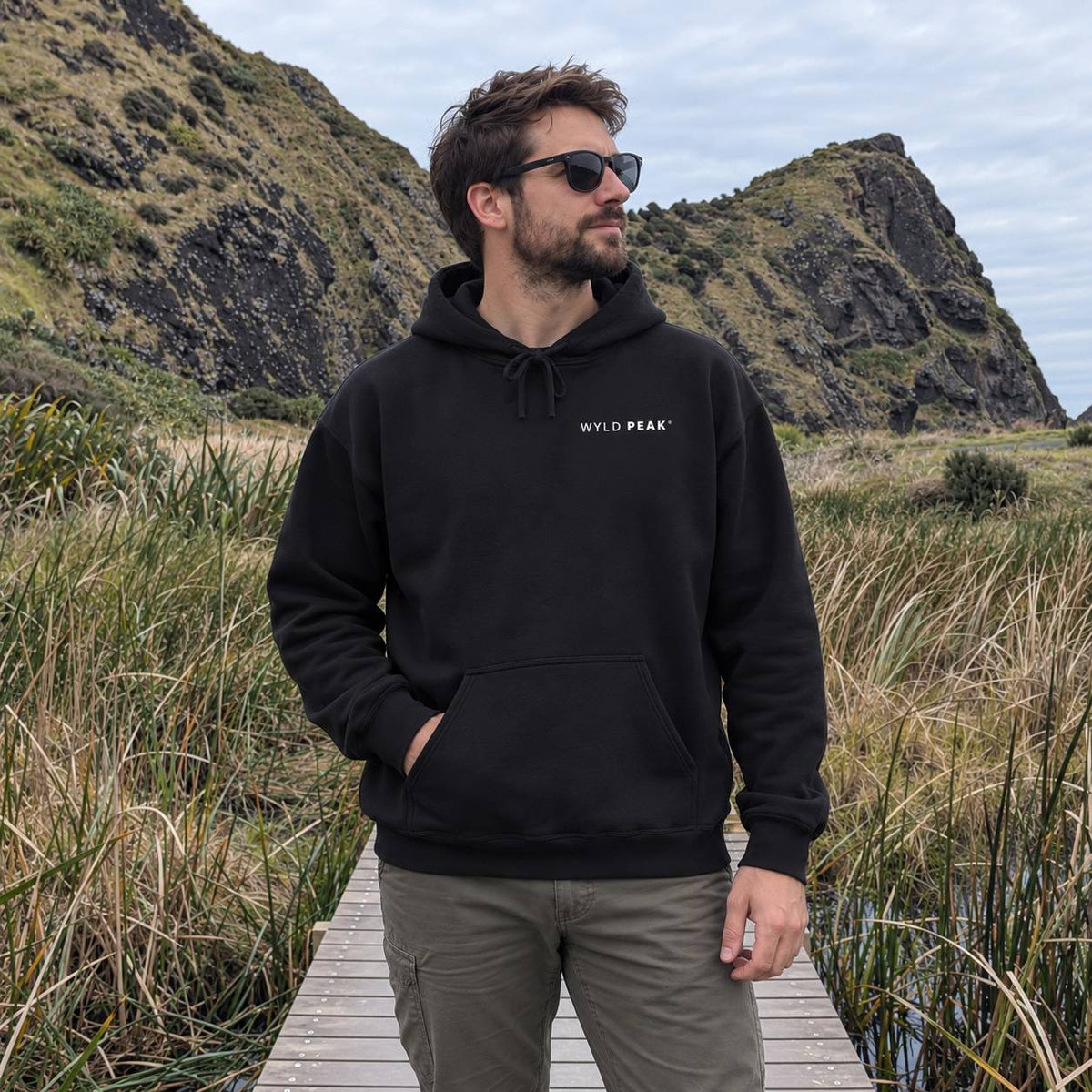 A man wearing a black pullover hoodie with 'WYLD PEAK' printed in white on the chest, standing on a wooden boardwalk in an outdoor grassy landscape with rocky hills in the background.