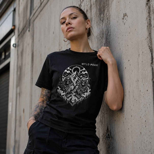 A woman wearing a black graphic t-shirt with a white circular mountain design and the text 'WYLD PEAK' on the chest, leaning against a concrete wall.