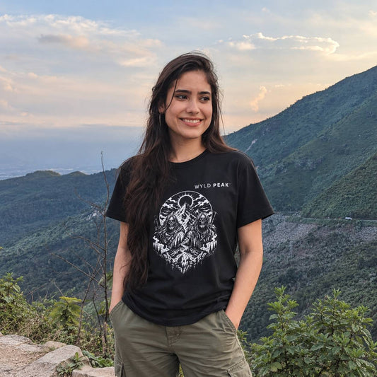 A woman stands outdoors in a mountain landscape wearing a black graphic T-shirt with a white circular wildcat design and the text 'WYLD PEAK' on the chest, paired with olive green cargo pants.