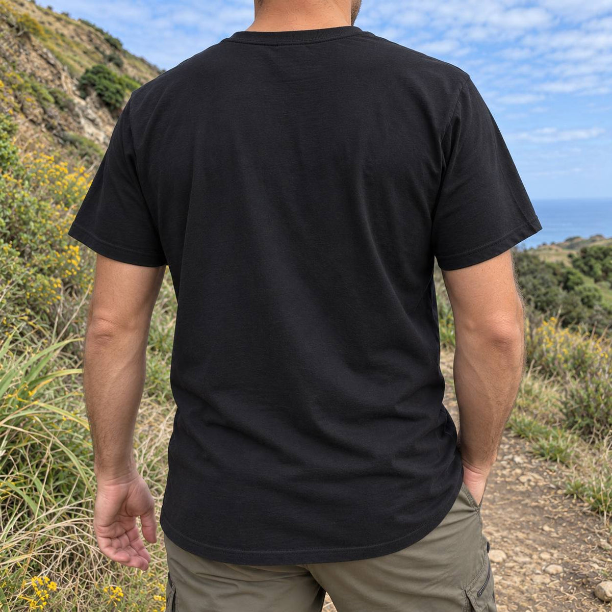 A man viewed from behind wearing a plain black short-sleeve t-shirt and olive green cargo pants, standing outdoors on a grassy hillside path with a blue sky and ocean in the background.