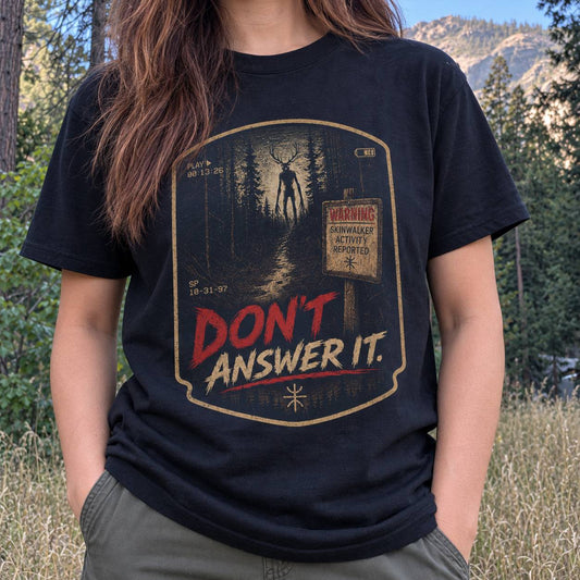 A person wearing a black graphic T-shirt outdoors in a forest setting. The shirt features a distressed vintage-style horror graphic with a deer-headed figure in a dark forest, a warning sign, and the text 'DON'T ANSWER IT.