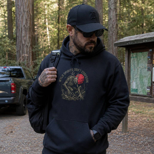 A man wearing a black hoodie with a graphic print that reads 'WILD SPIRITS DON'T CONFORM' and shows a red skull, walking in a forest setting.