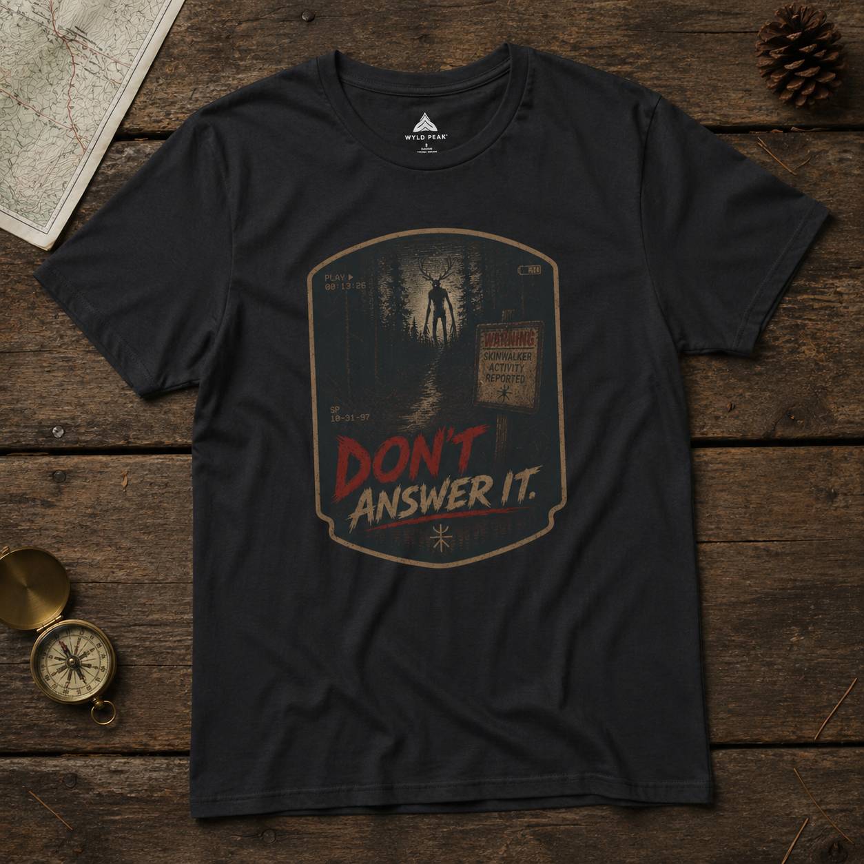 Black t-shirt with 'Don't Answer It'  skinwalker graphic on a wooden surface with map and compass.