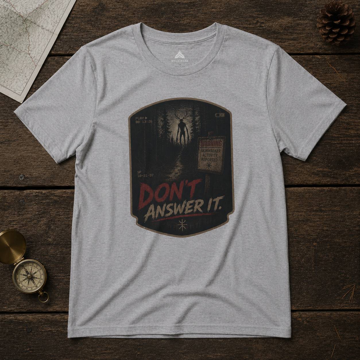 Gray t-shirt with 'Don't Answer It' skinwalker graphic design on a wooden surface with map and compass.
