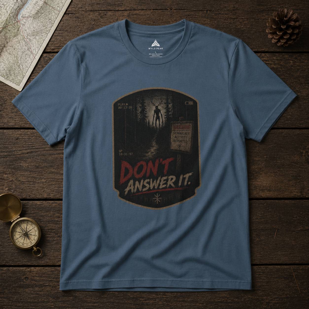Blue t-shirt with 'Don't Answer It' horror graphic on a wooden surface with map and compass.