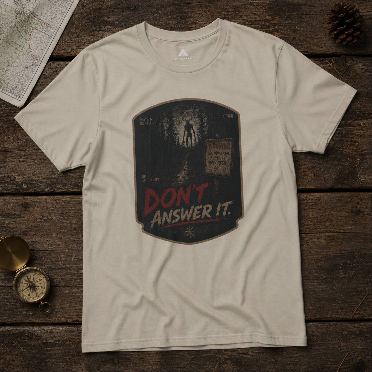 Beige t-shirt with 'Don't Answer It' skinwalker graphic on a wooden surface with map and compass.