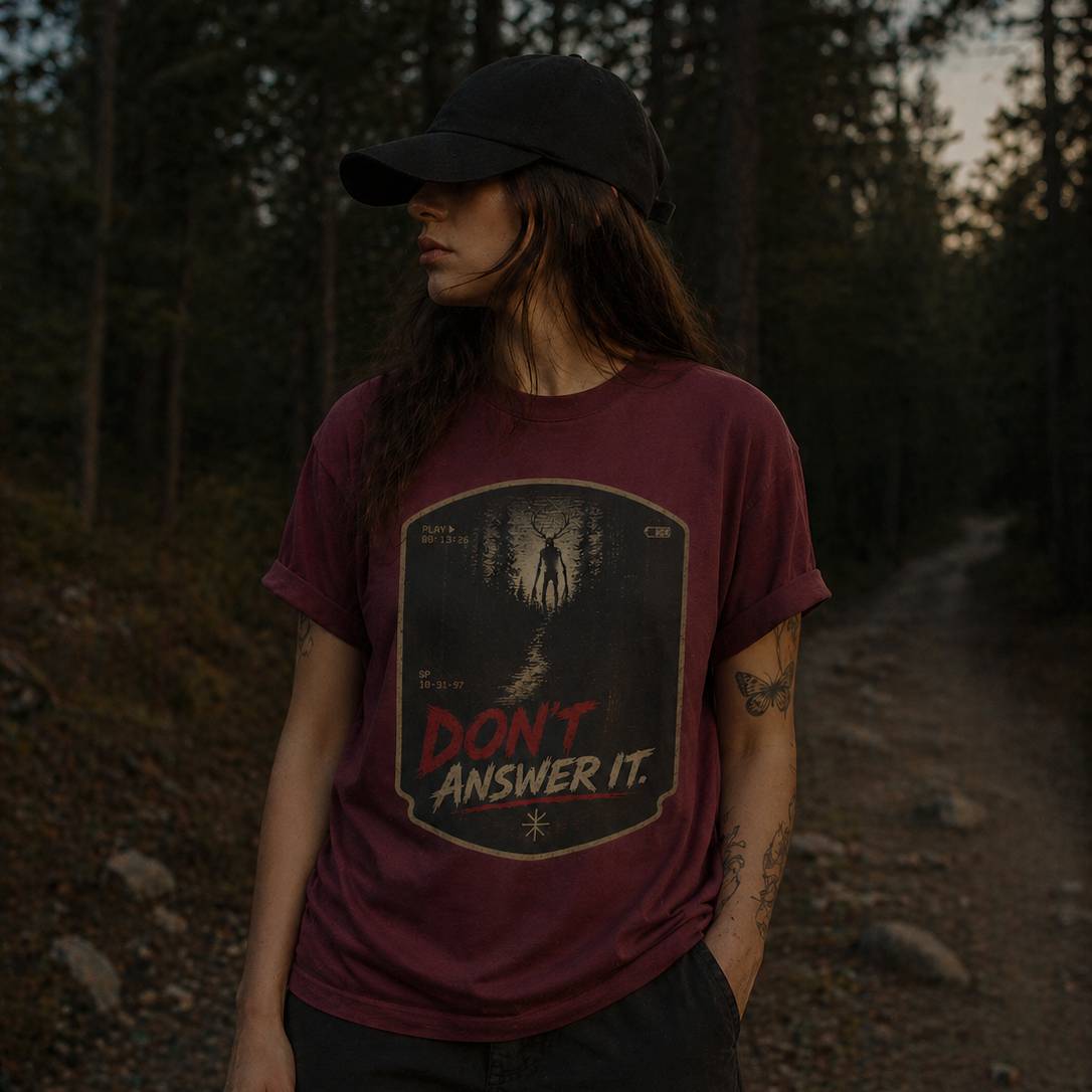 Person wearing a maroon t-shirt with a skinwalker cryptid graphic design and text in a forest setting