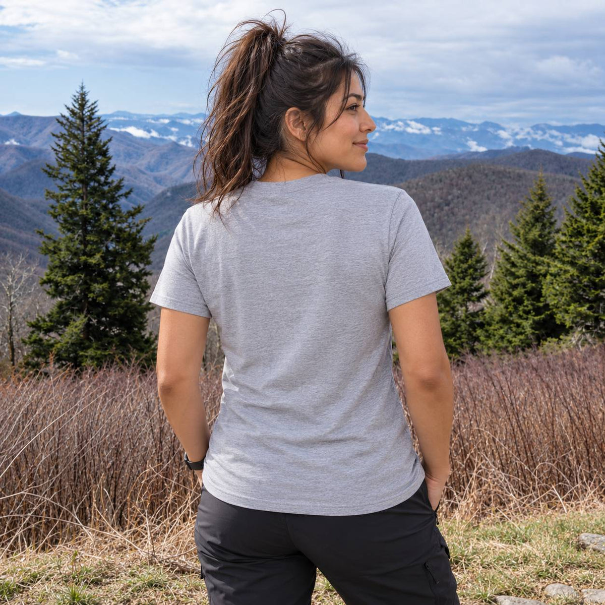 A woman wearing a light gray heather crewneck t-shirt and black cargo pants, photographed from behind as she looks out over a mountain landscape.