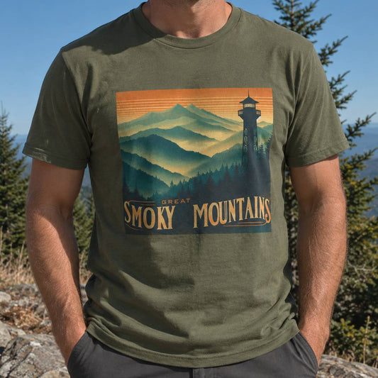 A man wearing an olive green t-shirt with a vintage-style graphic of the Great Smoky Mountains, set against a natural outdoor background of pine trees and mountains.