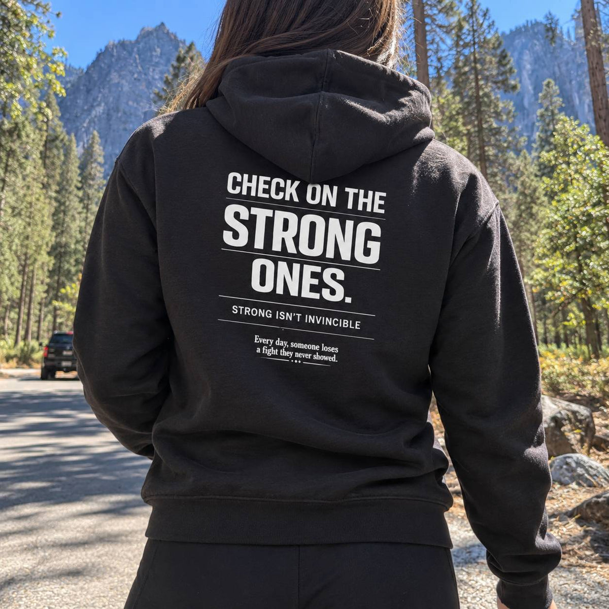 A person standing outdoors in a forest with mountains in the background, wearing a black hoodie from the back. The hoodie has large white text on the back that reads 'CHECK ON THE STRONG ONES.